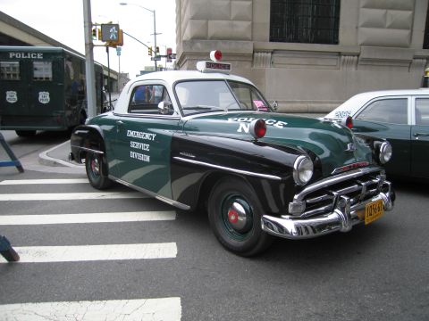 1991 Ford LTD Crown Victoria - restored green-and-black 1950s-style police coupe lettered 'Emergency Service Division' with a roof beacon at a city crosswalk.