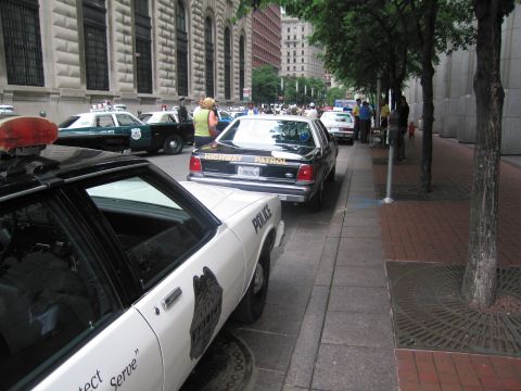 1991 Ford LTD Crown Victoria - mix of vintage and modern police cars, including a Highway Patrol sedan, lined along a downtown sidewalk with onlookers.