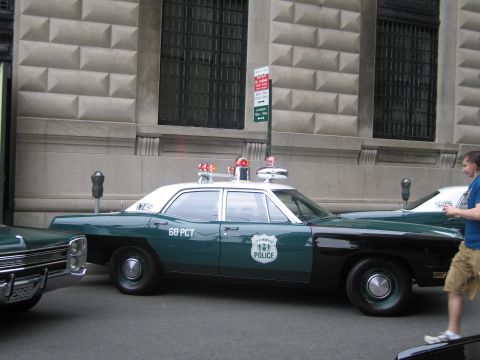 1991 Ford LTD Crown Victoria - restored green-and-black police sedan lettered '68 PCT' with red roof beacons parked beside a stone building, a man walking past.