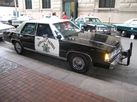 1991 Ford LTD Crown Victoria - black-and-white Ford Highway Patrol sedan with a gold star door shield and headlights on, parked on a brick sidewalk among other police car…