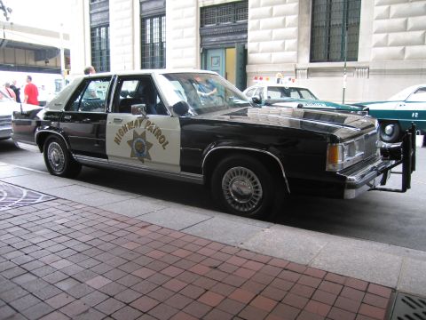 1991 Ford LTD Crown Victoria - black-and-white Highway Patrol sedan bearing a gold seven-point star door shield parked on a brick sidewalk near a stone building.