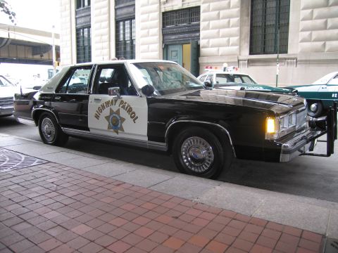 1991 Ford LTD Crown Victoria - front three-quarter view of a black-and-white Highway Patrol sedan with a gold star door shield and headlight on, parked on brick pavement.