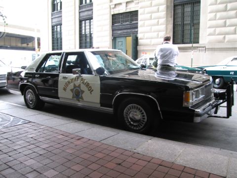 1991 Ford LTD Crown Victoria - black-and-white Highway Patrol sedan with a gold star door shield, a man in white kneeling near the hood, parked on a brick sidewalk.