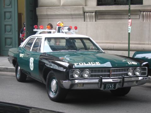 1991 Ford LTD Crown Victoria - restored green-and-white police sedan lettered 'POLICE 1745' with multiple roof beacons and a front 'NYC Police 1745' plate by a building.