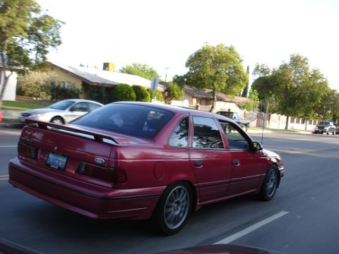 1993 Ford Taurus - A red 1993 Ford Taurus sedan is seen from the rear 3/4 angle, driving on a suburban street with other vehicles present.