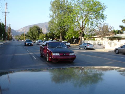1993 Ford Taurus - A red sedan with a black hood drives down a suburban street with mountains in the background.