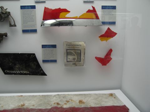 1991 Ford LTD Crown Victoria - museum case displaying recovered Crown Victoria police-car fragments, red and amber taillight pieces and printed placards on a white wall.