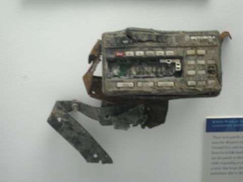 1991 Ford LTD Crown Victoria - close-up of a charred, dirt-covered Motorola handheld two-way radio with a bent bracket displayed on a white museum wall beside a placard.