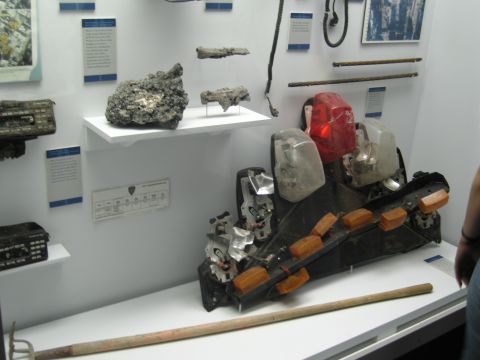 1991 Ford LTD Crown Victoria - crushed black plastic taillight housing with red and clear lenses sits on a museum exhibit shelf beside metal artifacts and wall placards.