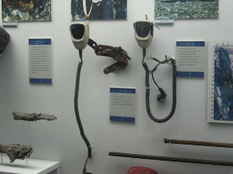 1991 Ford LTD Crown Victoria - Two worn microphone handsets with coiled cords hang on a white museum wall surrounded by photographs, placards, and metal relics.