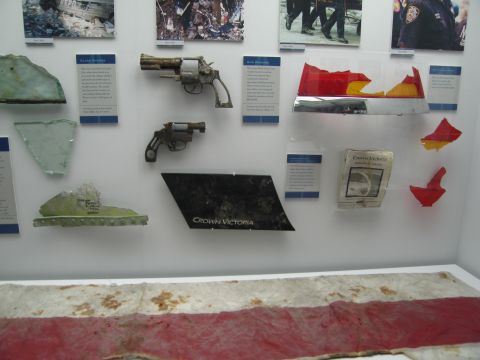 1991 Ford LTD Crown Victoria - exhibit wall shows two rusted revolvers, glass fragments, a black Crown Victoria badge panel, and red debris beneath documentary photos.
