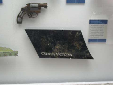 1991 Ford LTD Crown Victoria - rusted revolver hangs above a black panel bearing the chrome Crown Victoria nameplate, displayed on a white museum wall with a placard.