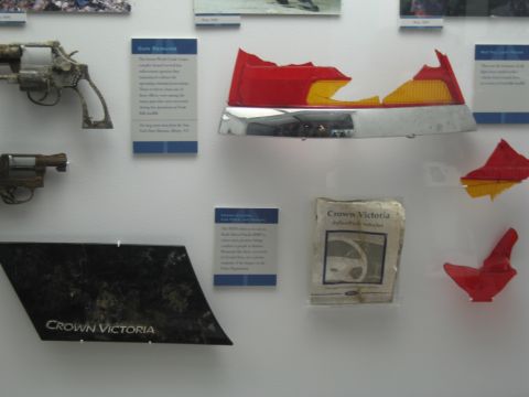 1991 Ford LTD Crown Victoria - museum wall displays rusted revolvers, twisted red and chrome trim, an owner's manual, and a black Crown Victoria emblem panel.