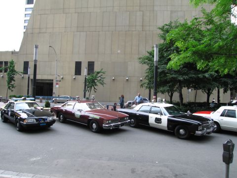 1991 Ford LTD Crown Victoria - Classic police and patrol cars, including black-and-white sedans and a maroon coupe, line a city curb beside a large stone building.