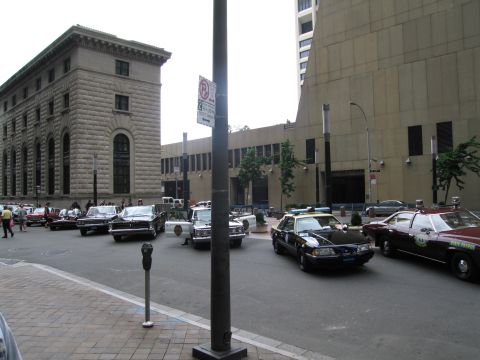 1991 Ford LTD Crown Victoria - city street scene shows several vintage police cars parked and arrayed near tall stone and concrete buildings behind a lamp post.