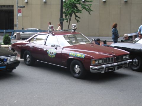 1991 Ford LTD Crown Victoria - maroon classic four-door State Patrol sedan with a roof beacon and door decals sits among other vintage cars on a city street.