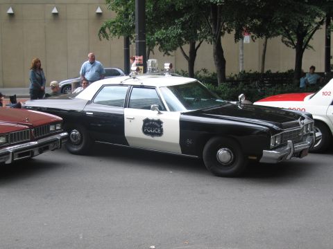1991 Ford LTD Crown Victoria - black-and-white classic Police sedan with a roof spotlight and door shield emblem is parked among other vintage cars near trees.