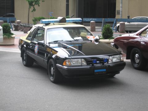 1991 Ford LTD Crown Victoria - black State Trooper patrol coupe with a roof light bar and Ford badge, resembling a Fox-body Mustang, sits parked on a city street.