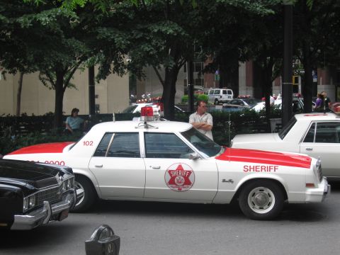 1991 Ford LTD Crown Victoria - white and red classic Sheriff sedan marked 102 with a roof beacon and star emblem is parked along a tree-lined city street.