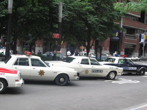 1991 Ford LTD Crown Victoria - Several vintage Sheriff and Police sedans with roof beacons line a city curb beneath trees in front of brick buildings.
