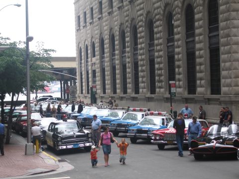 1991 Ford LTD Crown Victoria - People and children walk among a long row of vintage police cars, including a black car, parked beside a large arched stone building.