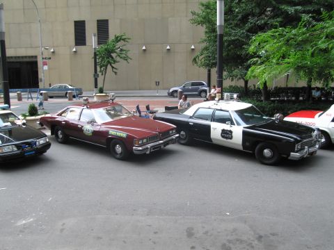 1991 Ford LTD Crown Victoria - maroon State Patrol sedan sits beside a black-and-white Police sedan, both with roof beacons, along a city curb near a concrete building.