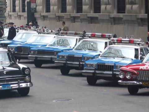 1991 Ford LTD Crown Victoria - line of blue-and-white vintage police sedans with roof beacons sits angle-parked along a city street near older black cars.