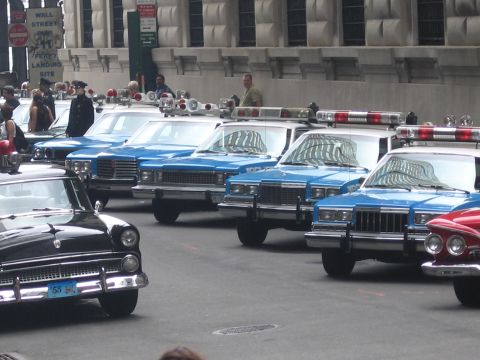 1991 Ford LTD Crown Victoria - Blue-and-white vintage police sedans with roof beacons sit angle-parked near a Wall Street ferry landing sign and older black cars.
