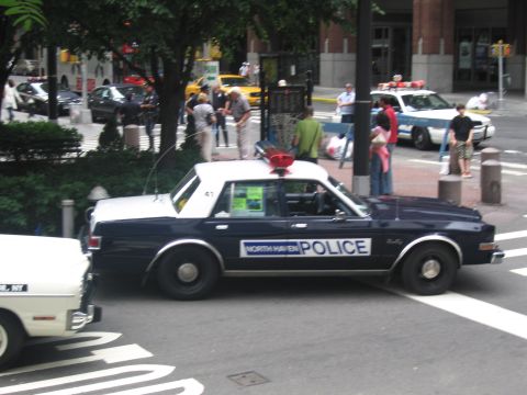 1991 Ford LTD Crown Victoria - dark North Haven Police black-and-white sedan with a roof spotlight maneuvers through a city intersection past pedestrians.