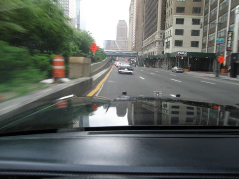 1991 Ford LTD Crown Victoria - Seen over a car's dashboard and hood, a white police-style car drives ahead down a city avenue lined with skyscrapers and a PARK sign.