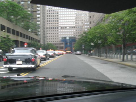 1991 Ford LTD Crown Victoria - From inside a car, a black vintage sedan with round taillights drives ahead on a city street flanked by tall buildings and trees.