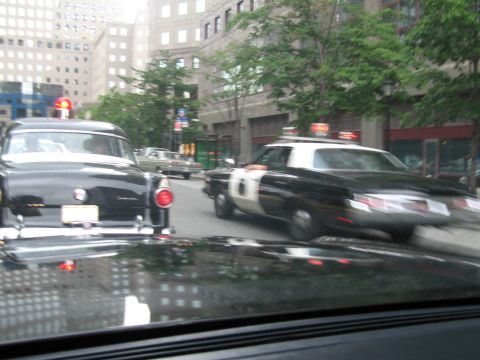 1991 Ford LTD Crown Victoria - Through a windshield, a black-and-white police car crosses an intersection beside a black vintage sedan amid downtown buildings.