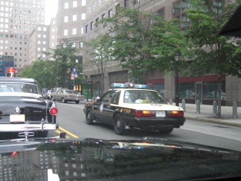 1991 Ford LTD Crown Victoria - Seen through a windshield, a black-and-tan police sedan with a roof light drives on a downtown street lined with skyscrapers and trees.