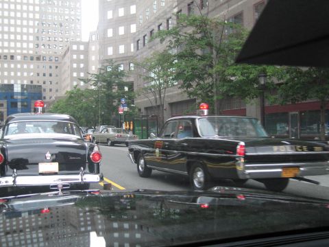 1991 Ford LTD Crown Victoria - Through a windshield, a black vintage Police sedan with a red roof beacon drives down a city avenue past other classic cars and skyscrapers.