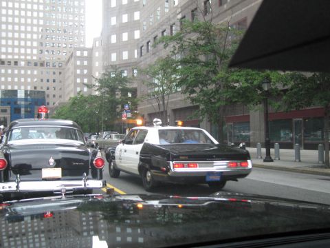 1991 Ford LTD Crown Victoria - black-and-white vintage police sedan with a roof beacon drives down a city street past parked classic cars amid tall buildings.
