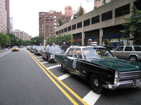 1991 Ford LTD Crown Victoria - Several green-and-white classic police sedans are lined up along a city curb beside a parking garage, with a yellow taxi passing in the bac…