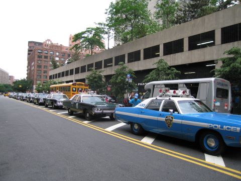 1991 Ford LTD Crown Victoria - blue-and-white classic Police sedan parks at a city curb leading a row of vintage police vehicles, a yellow school bus and a paddy-wagon vi…