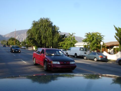 1993 Ford Taurus - A red sedan with a black hood drives down a suburban street with mountains in the background.