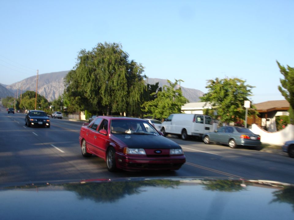 Red sedan with a black hood driving on a street, front view. The car appears to be from the early 1990s