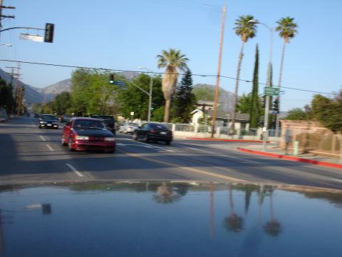 1993 Ford Taurus - A red sedan with a black hood drives down a suburban street with palm trees and mountains visible.