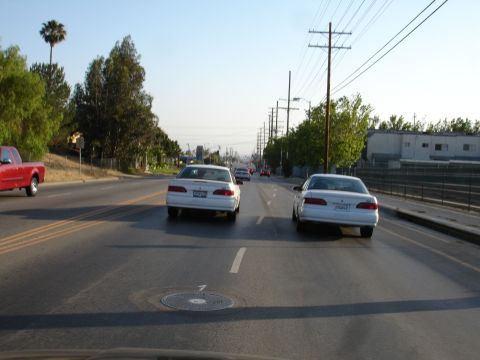 1993 Ford Taurus - Two white sedans are seen from behind on a sunny day, driving down a multi-lane road with power lines overhead.