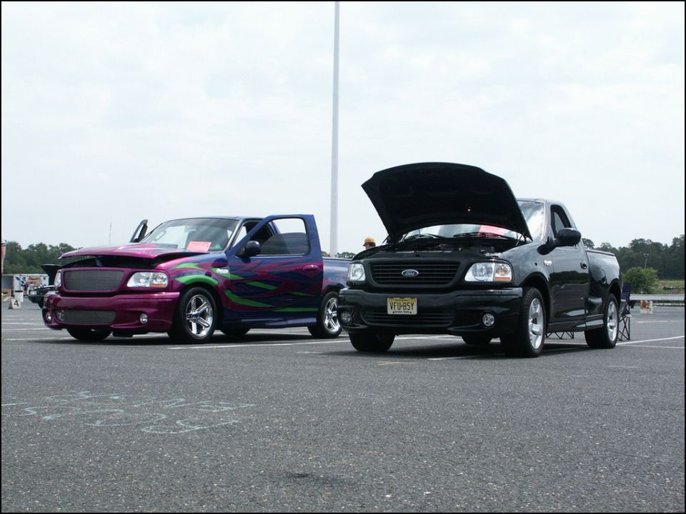 Two Ford F-150 Lightning trucks, one purple with custom graphics and one black with hood open, parked on asphalt