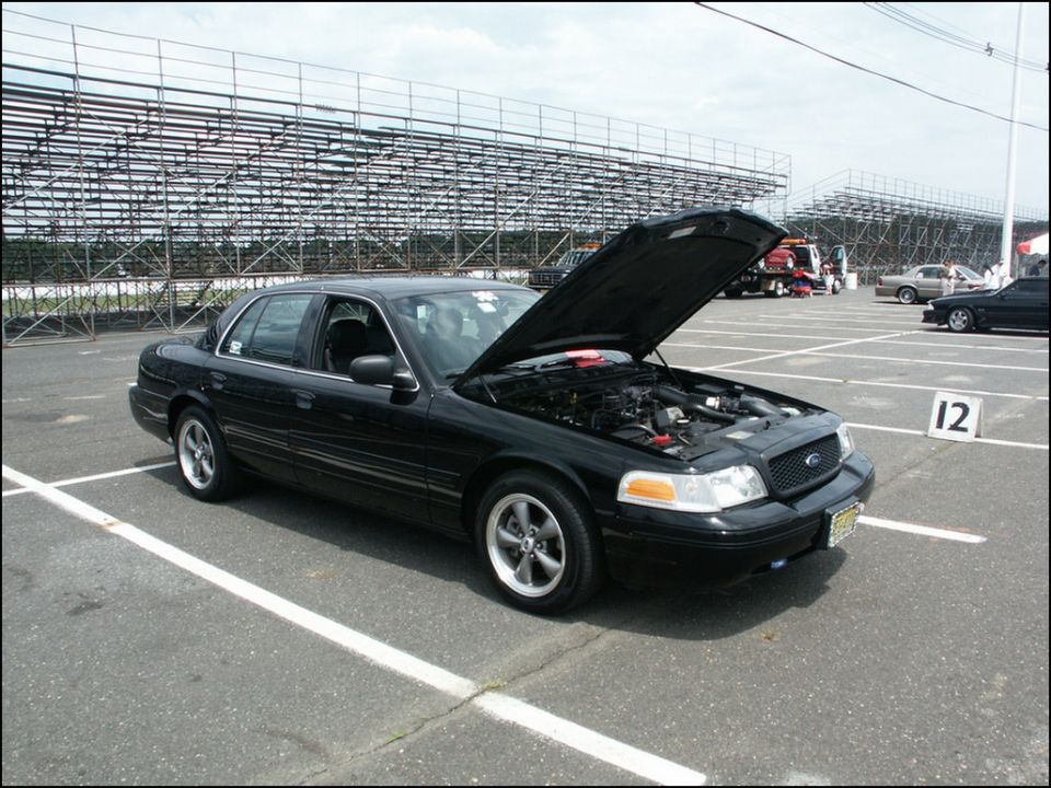 Black sedan with hood open, parked in a lot with bleachers in the background. Another car is visible to the right