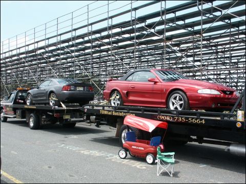 2001 Mercury Grand Marquis - Two Ford Mustangs, a red convertible and a gray coupe, are transported on a tow truck in front of stadium bleachers.