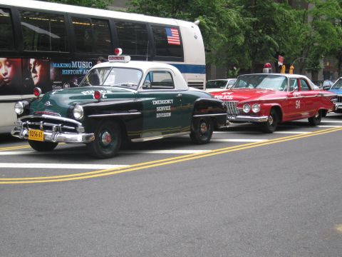 1991 Ford LTD Crown Victoria - dark green vintage Emergency Service Division police coupe sits next to a red police car on a city street, with a bus and crowd behind.