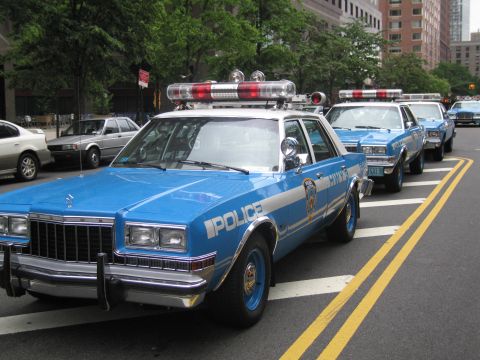 1991 Ford LTD Crown Victoria - blue-and-silver POLICE sedan with a red roof lightbar parks at a city curb, leading a line of matching vintage patrol cars.