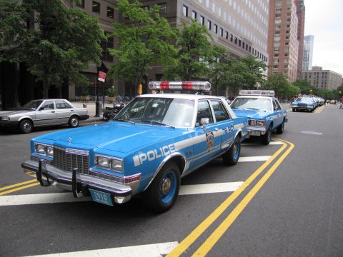 1991 Ford LTD Crown Victoria - blue POLICE sedan with a red roof lightbar parks at a city curb, with additional vintage patrol cars lined up behind it.