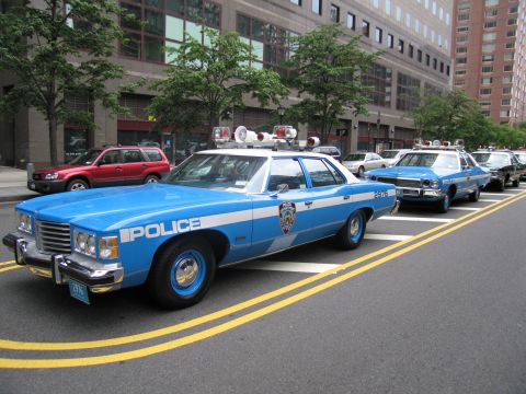 1991 Ford LTD Crown Victoria - light blue POLICE sedan with a roof-mounted speaker parks at a city curb leading a row of vintage patrol cars, a red SUV passing nearby.