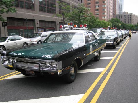 1991 Ford LTD Crown Victoria - dark green-and-white POLICE sedan numbered 2902 with a red roof beacon parks at a city curb, leading a line of vintage patrol cars.