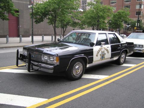1991 Ford LTD Crown Victoria - black-and-white Highway Patrol sedan fitted with a front push bar and gold star parks at a city crosswalk beside another patrol car.